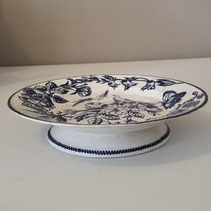 Pode incluir: Uma taça redonda e rasa sobre pedestal com base branca e um padrão de transferência azul escuro. O interior apresenta ilustrações detalhadas de flores, folhas e um pássaro. A borda e a base são decoradas com uma fina borda pontilhada.