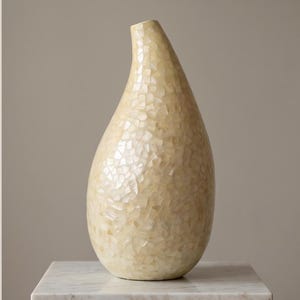 Peut inclure: Un vase en forme de goutte d'eau avec un col effilé et une base large et arrondie. La surface extérieure est recouverte d'une mosaïque de petits fragments de coquillages irisés de couleur crème, créant un aspect texturé et réfléchissant.