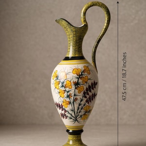 Peut inclure: Une grande cruche en céramique avec un col, une anse et une base vert olive mouchetés. Le corps blanc présente un motif botanique peint à la main avec des fleurs jaunes et un feuillage sombre. Une ligne de mesure indique 47,5 cm de hauteur.