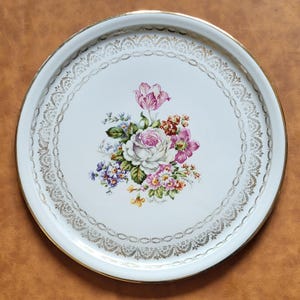 Peut inclure: Un plateau de service rond blanc avec un bord doré et un motif floral. Le plateau présente un bouquet central de fleurs roses, violettes et blanches. Un motif de filigrane doré entoure l'arrangement floral et le bord extérieur.