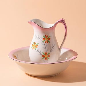 Può includere: Un set composto da una bacinella e una brocca in ceramica bianca con bordo rosa e un motivo floreale di fiori arancioni e verdi con steli grigi. La brocca è dotata di un manico curvo e di un beccuccio, la bacinella ha una forma ampia e bassa.
