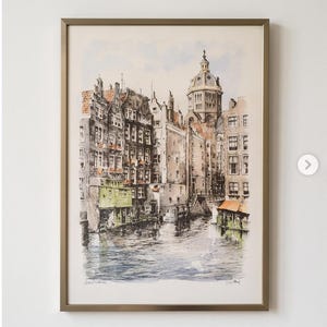 Könnte beinhalten: Ein gerahmtes Aquarellbild, das eine Kanalszene in Amsterdam mit hohen, schmalen Gebäuden, einem markanten Kirchturm mit Kuppel und Spiegelungen auf dem Wasser zeigt. Das Kunstwerk ist in Grau-, Braun- und Orangetönen gehalten und trägt den Text Amsterdam.