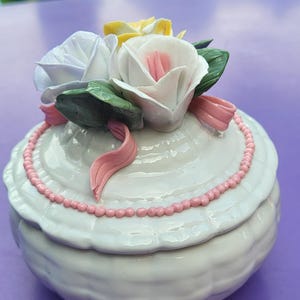 Peut inclure: Une boîte à bijoux en céramique blanche avec une texture tressée. Le couvercle présente un bouquet de roses sculptées en blanc, jaune et rose, agrémenté de feuilles vertes et d'un ruban rose. Une fine bordure perlée rose entoure le couvercle.
