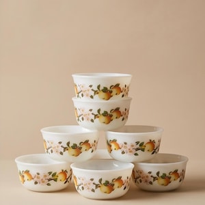 Peut inclure: Une pile de six bols en verre blanc disposés en pyramide. Chaque bol présente un motif répétitif de pommes jaunes, de feuilles vertes et de fleurs rose clair entourant l'extérieur. Les bols ont un bord et une base arrondis et lisses.