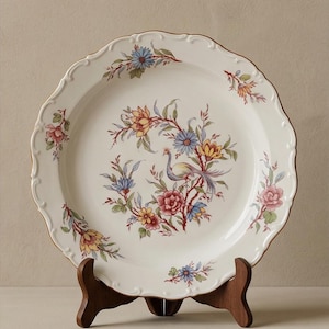 May include: A round white ceramic plate with a scalloped edge and a gold-toned rim. The center features a colorful illustration of a bird surrounded by various flowers in shades of blue, yellow, pink, and green. Additional floral motifs are placed along the rim.
