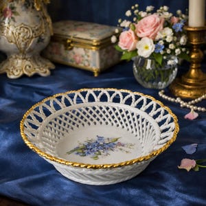 Peut inclure: Un bol en porcelaine blanche doté d'un bord ajouré en forme de treillis avec une bordure dorée en corde torsadée. Le centre du bol est orné d'un motif floral peint composé de myosotis bleus et de feuilles vertes sur un fond blanc.