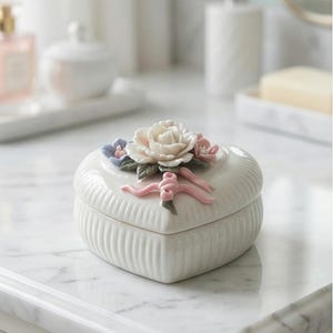 Peut inclure: Une boîte à bijoux en céramique blanche en forme de cœur avec des côtés côtelés verticaux. Le couvercle présente un arrangement floral sculpté avec une grande rose blanche centrale, des fleurs roses et bleues, des feuilles et un ruban rose.