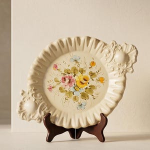 Peut inclure: Une assiette en céramique couleur crème avec un bord festonné et des détails en relief sur deux côtés. Le centre présente un motif floral peint à la main avec des fleurs roses, jaunes et bleu clair entourées de feuilles vertes et de tiges brunes.
