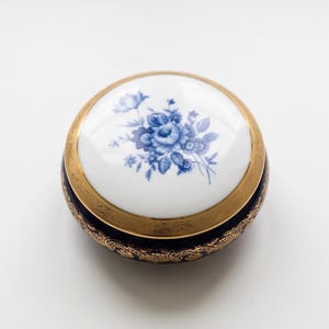Può includere: Un portagioie rotondo in porcellana con una base blu navy profondo e intricati motivi in filigrana dorata. Il coperchio è bianco con un'illustrazione centrale di un bouquet di fiori blu e un ampio bordo dorato con dettagli incisi.