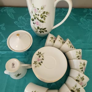 Puede incluir: Un juego de té de porcelana blanca que incluye una tetera alta con tapa, un azucarero con tapa, una lechera y varias tazas con sus platos a juego. Cada pieza está decorada con un delicado patrón de rosas rosadas y hojas verdes con bordes dorados.