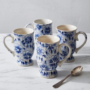 Peut inclure: Un ensemble de trois tasses en céramique avec une base blanche et un motif floral bleu, chacune dotée d'un corps arrondi, d'une anse incurvée et d'une base sur pied. Une cuillère en métal argenté est placée à droite des tasses sur la surface.