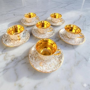 Peut inclure: Un ensemble de six tasses à thé et soucoupes en porcelaine avec une base blanche, des motifs floraux dorés complexes et un intérieur doré réfléchissant. Chaque tasse possède une soucoupe assortie avec un bord doré et un motif botanique.