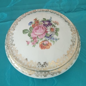 Pode incluir: Uma caixa de joias redonda de porcelana branca com uma tampa apresentando um buquê floral central de rosas cor-de-rosa, papoulas laranja e flores roxas. A tampa e a base são decoradas com um intrincado padrão de borda de filigrana dourada.