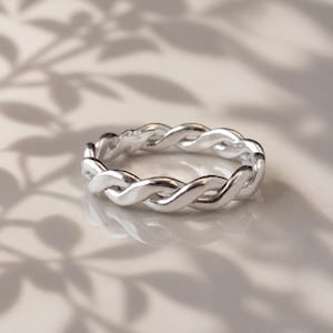 Puede incluir: Un anillo plateado con un diseño trenzado. El anillo es circular y presenta un patrón continuo de hebras entrelazadas. El anillo se muestra sobre una superficie clara con sombras de hojas.