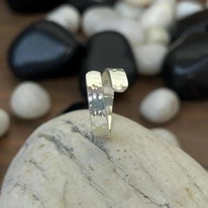 May include: A silver-colored ring with a hammered texture. The ring has a unique wrap-around design, with one end curving over the top. The ring is displayed on a light-colored stone, with black and white stones in the background.