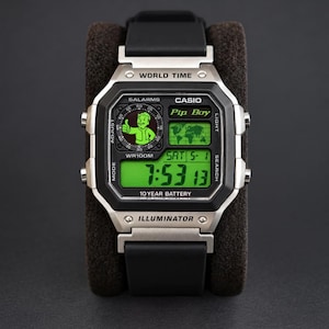 May include: A digital Casio World Time watch with a black resin strap and a silver-tone rectangular case. The display features a green background with a Pip Boy graphic, a world map, and the text "Pip Boy," "5 ALARMS," "WR100M," and "10 YEAR BATTERY."
