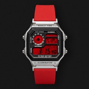 Könnte beinhalten: Eine digitale Armbanduhr mit silbernem Gehäuse und rotem Armband. Das Zifferblatt zeigt die Uhrzeit in roten Ziffern an. Auf dem Zifferblatt der Uhr steht das Wort "CASIO". Die Uhr ist vor einem schwarzen Hintergrund platziert.