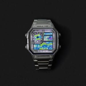 Op de afbeelding: Een zilveren Casio digitale horloge met een rechthoekige wijzerplaat en een bijpassende roestvrijstalen band. De wijzerplaat geeft de tijd, datum en andere functies weer. Het horloge heeft de tekst "WORLD TIME", "CASIO" en "10 YEAR BATTERY".