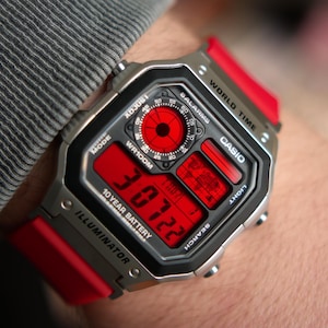 以下が含まれることがあります： 赤色のディスプレイとバンドが付いた、シルバーのCasioデジタル腕時計。文字盤には時刻、日付、その他の機能が表示されています。時計には「WR100M」、「10 YEAR BATTERY」、「ILLUMINATOR」の文字があります。