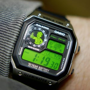 May include: A digital Casio watch with a black and silver case, displaying the time in green digits. The watch face features a Vault Boy graphic and the text "WR100M". The watch is on a wrist.