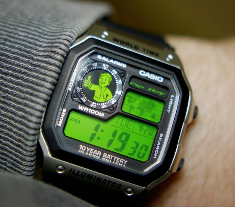 May include: A digital Casio watch with a black and silver case, displaying the time in green digits. The watch face features a Vault Boy graphic and the text "WR100M". The watch is on a wrist.