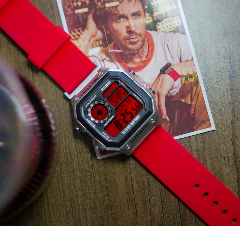 May include: A Casio digital watch with a red strap and a black and silver face. The watch displays the time in red digits. The watch is on a wooden surface, with a picture of a man in the background.