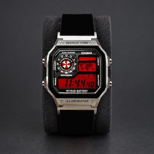 May include: A digital wristwatch with a silver-tone rectangular case and a black strap. The watch face features a red display with the text CASIO, WORLD TIME, and ILLUMINATOR. A circular graphic with the Umbrella Corporation logo is visible.