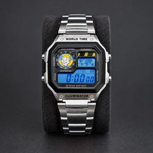 May include: A silver-tone digital wristwatch with a stainless steel link band. The square face features a black bezel, a blue digital display, and a graphic of a cartoon character in a yellow jumpsuit. Text includes WORLD TIME, CASIO, and 10 YEAR BATTERY.