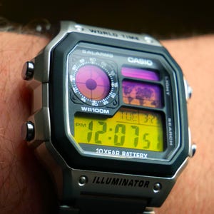 Op de afbeelding: Een zilveren en zwarte Casio digitale horloge met 12:07 PM. De wijzerplaat heeft een gele digitale display, een wereldkaart en de tekst "WR100M" en "ILLUMINATOR". Het horloge zit om een pols.
