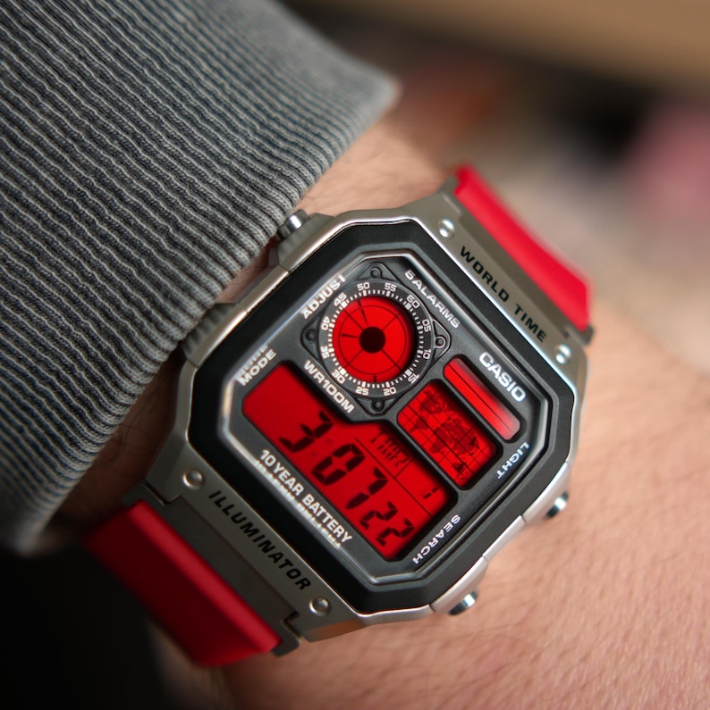 以下が含まれることがあります： 赤色のディスプレイとバンドが付いた、シルバーのCasioデジタル腕時計。文字盤には時刻、日付、その他の機能が表示されています。時計には「WR100M」、「10 YEAR BATTERY」、「ILLUMINATOR」の文字があります。