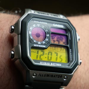 Puede incluir: Un reloj digital Casio plateado y negro con una pantalla digital amarilla que muestra la hora. La esfera del reloj incluye un mapa del mundo y el texto "WORLD TIME", "BALARMS", "WR100M" e "ILLUMINATOR".