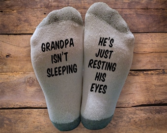 May include: A pair of white socks with black text. One sock reads "Grandpa isn't sleeping" and the other sock reads "He's just resting his eyes".