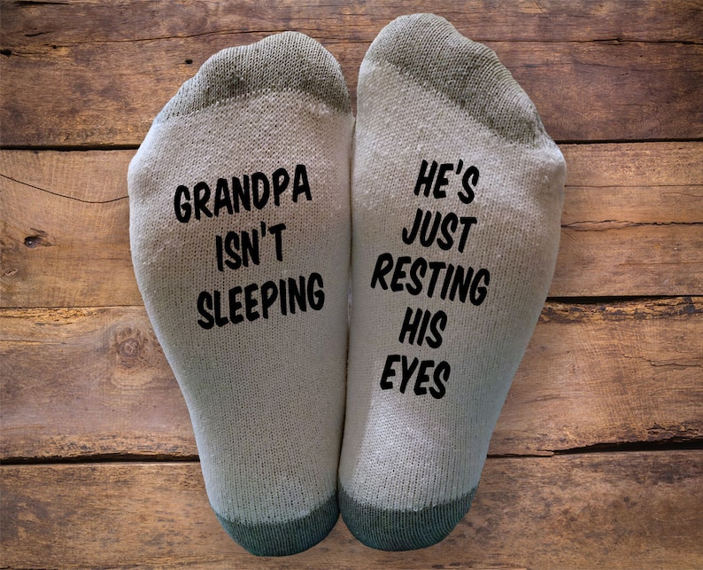 Op de afbeelding: Een paar witte sokken met zwarte tekst. E&eacute;n sok heeft de tekst "Grandpa isn't sleeping" en de andere sok heeft de tekst "He's just resting his eyes".