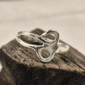 May include: A silver ring with an unusual open design. The ring has a central figure that looks like a figure eight or an infinity symbol. The ring is displayed on a piece of dark wood.