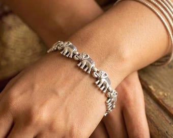 May include: Silver-toned elephant bracelet. The bracelet features a series of small, detailed elephant charms linked together. The elephants have textured details and are arranged in a row. The bracelet is worn on a wrist, with other silver bracelets visible.