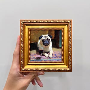 May include: A framed painting of a pug dog with a glass of red wine. The pug is white and black, lying on a patterned rug. The frame is gold with ornate detailing. The painting has a vintage aesthetic.
