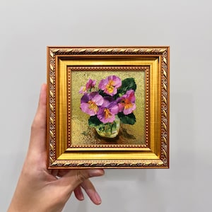 以下が含まれることがあります： 装飾的なゴールドのフレームに入った正方形の油絵。紫と黄色のパンジーが活けられた小さなガラスの花瓶が描かれています。テクスチャのあるゴールドの背景が、鮮やかな花の色と華やかなフレームのディテールを際立たせています。