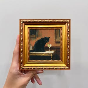May include: A framed painting depicts a black cat sitting on a table with a martini glass. The painting has a gold frame with ornate detailing. The cat has yellow eyes and is looking at the martini.