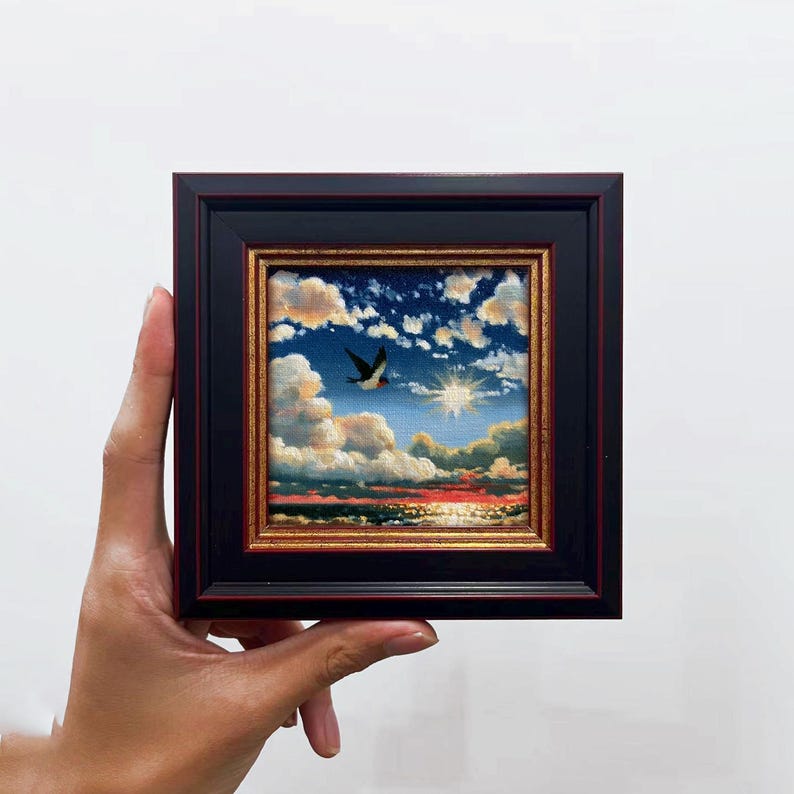 May include: A small square oil painting in a black frame with gold inner trim. The artwork depicts a sunset over the ocean with a single bird flying through a blue sky filled with white and orange clouds and a bright sunburst in the center.