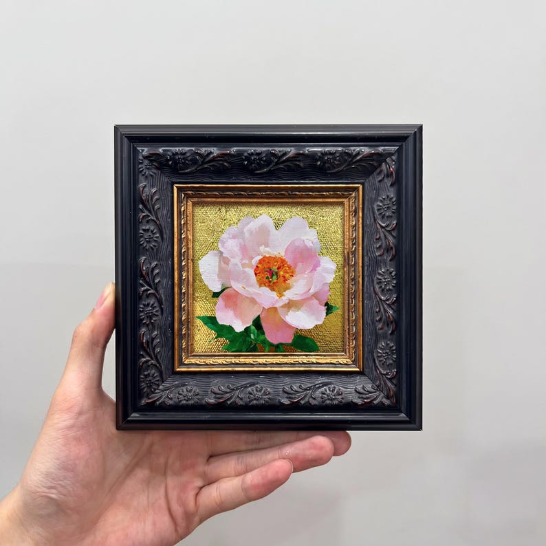 Op de afbeelding: Een klein vierkant schilderij van een lichtroze pioenroos met een feloranje hart, tegen een getextureerde gouden achtergrond. Het kunstwerk zit in een donkere, sierlijke, gesneden houten lijst met gedetailleerd bloemreli&euml;f.