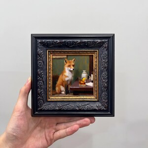 May include: A framed painting of a fox at a table with a drink, set within a black ornate frame with a gold inner border. The fox has orange and white fur, and the drink has a lemon garnish. The background is a muted green, creating a classic aesthetic.