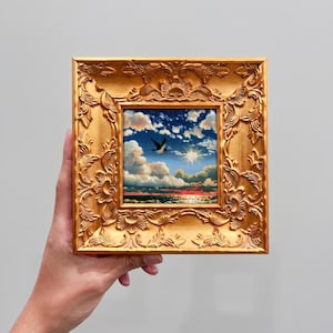 May include: A square painting in an ornate gold frame featuring a sunset over the ocean with fluffy clouds and a single bird in flight. The sky transitions from deep blue to warm orange hues near the horizon, reflecting light on the water surface.
