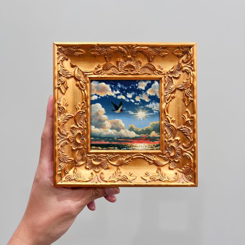 May include: A square painting in an ornate gold frame featuring a sunset over the ocean with fluffy clouds and a single bird in flight. The sky transitions from deep blue to warm orange hues near the horizon, reflecting light on the water surface.
