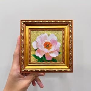 Op de afbeelding: Een vierkant schilderij van een lichtroze pioenroos met een feloranje hart en groene bladeren, tegen een getextureerde gouden achtergrond. Het kunstwerk zit in een sierlijke, metallic gouden lijst met een decoratief bladpatroon.