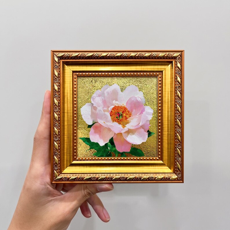Op de afbeelding: Een vierkant schilderij van een lichtroze pioenroos met een feloranje hart en groene bladeren, tegen een getextureerde gouden achtergrond. Het kunstwerk zit in een sierlijke, metallic gouden lijst met een decoratief bladpatroon.