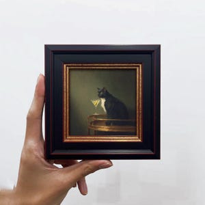 May include: A framed painting depicts a black and white cat sitting next to a cocktail. The painting has a gold and black frame. The cat is sitting on a gold table, and the background is a muted green.