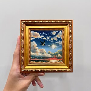 May include: A small square painting in an ornate gold frame featuring a landscape scene of a bird flying across a blue sky with white clouds, a bright sun, and a red sunset horizon over a dark sea. The frame has a textured, decorative border.
