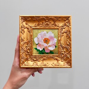 Op de afbeelding: Een vierkant olieverfschilderij van een enkele lichtroze pioenroos met een feloranje hart en groene bladeren, tegen een getextureerde gouden achtergrond. Het kunstwerk zit in een sierlijke, metallic gouden lijst met bloemmotieven.