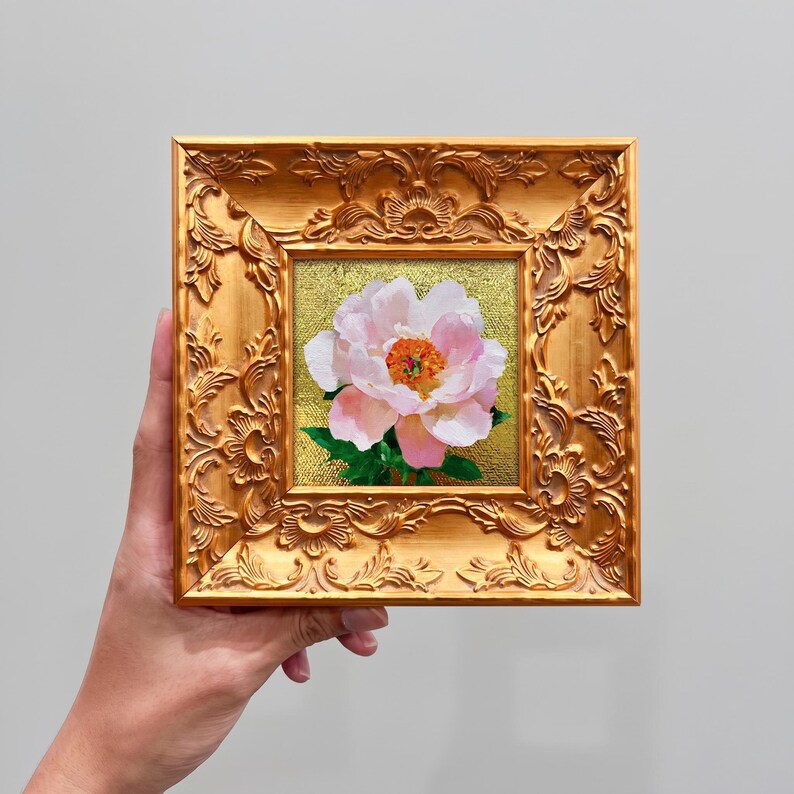 Op de afbeelding: Een vierkant olieverfschilderij van een enkele lichtroze pioenroos met een feloranje hart en groene bladeren, tegen een getextureerde gouden achtergrond. Het kunstwerk zit in een sierlijke, metallic gouden lijst met bloemmotieven.