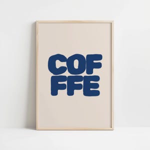 Op de afbeelding: Een ingelijste print met het woord "COFFEE" in vette, donkerblauwe letters. De letters zijn gestapeld, met "COF" boven en "FFE" eronder, tegen een lichtbeige achtergrond. De lijst heeft een lichte houtkleur.